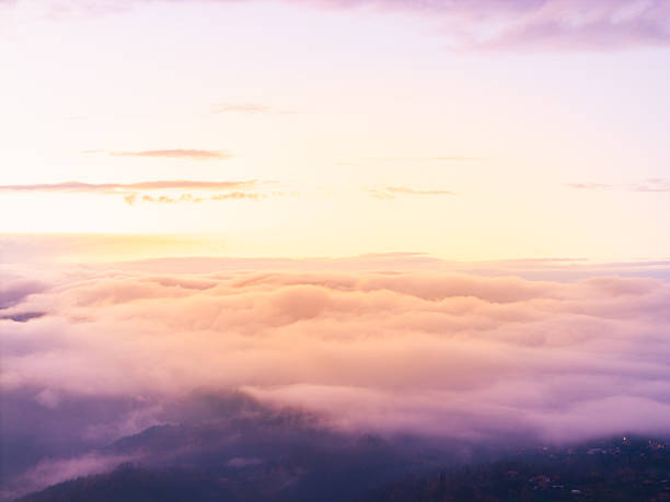 Drone image capturing the sun sinking above wide clouds and drifting fog. Abstract, minimal and atmospheric view showcasing soft light gradients, calm mood and dreamy horizon.