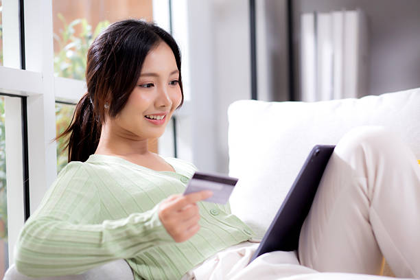 istock Young asian woman sitting on sofa using tablet computer shopping online with credit card buying to internet while cheerful, female payment with business, purchase and payment, business and finance. 2248490977