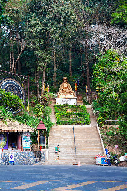 Golden thai monk statue above steps near entrance to Wat Doi Suthep in Chiang Mai. Statue is Kruba Srivichai, The Golden Monk, who was engineer for rebuilding temples. A thai man is sitting on steps below and is watching mobile phone, in front of him is a dog. At right side are some femalew thai street food vendors.
