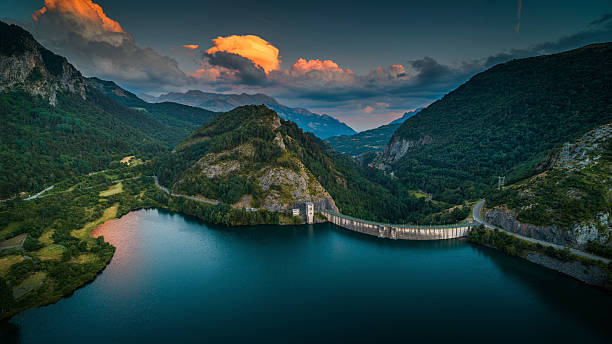 Aerial view of the Embalse de Lanuza in the Pyrenees of Huesca, Aragón, Spain, formed by the Río Gállego. A curved concrete dam stretches across the deep blue water, embraced by steep mountains and rocky slopes. Above, soft orange and pink clouds glow in the fading light, standing out against a deepening blue sky. The layers of mountains in the distance fade into the horizon. The calm surface of the reservoir and the dramatic light in the sky come together to create a peaceful yet powerful mountain scene. Lanuza in the municipality of Sallent de Gállego, in the province of Huesca, Aragon - Europe.