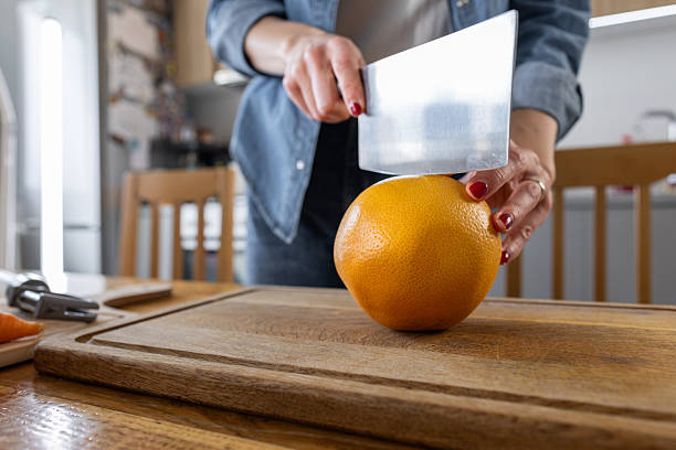 A person in a denim shirt slices a bright orange with a large blade over a wooden cutting board in a clean, well-lit kitchen. Fresh fruit and cooking moment captured.