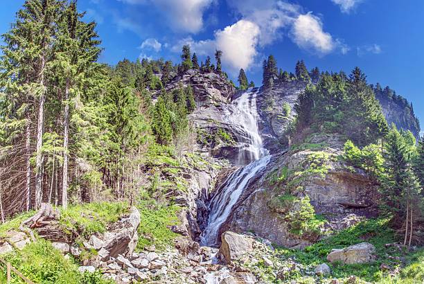 Wide panorama featuring a tall alpine waterfall flowing down rocky mountain slopes surrounded by dense evergreen forest.