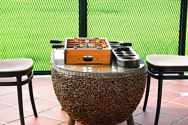 Small wicker table with a foosball game and two wooden chairs on a tiled patio, enclosed by a mesh railing with a scenic view of green fields and trees.