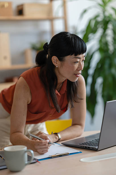 istock Asian woman small entrepreneur leaning over her desk in a bright home office, taking notes while working on her laptop with focus and determination 2248513193