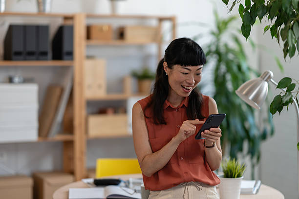 istock Asian woman small entrepreneur checking her smartphone in a bright home office, smiling while managing her business tasks with focus and energy. 2248516257