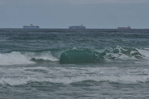 istock Waves crash on a beach with cargo ships visible in the distance on a cloudy day 2248524757