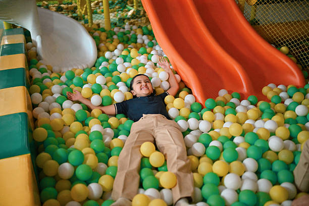 A cheerful young girl student in uniform lies on her back in a colorful ball pit at an indoor playground, smiling with joy and stretching her arms. Enjoying a moment of carefree fun near slides. Childhood happiness and playful energy in a safe and engaging environment.