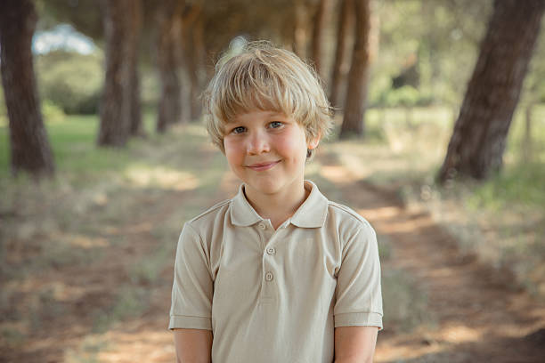 Smiling blond boy standing outdoors in a forest, natural light portrait, happy child looking at the camera.