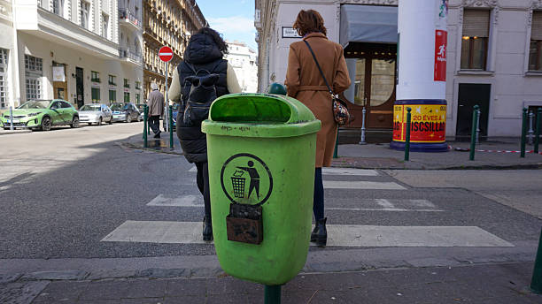 Budapest, Hungary 08-03-2025. Vibrant urban scene featuring a bright green trash bin in the foreground as people walk across a crosswalk. Waste managment in the city, encouraging to avoid littering
