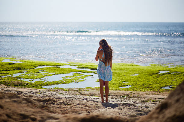 Girl standing on sandy beach facing ocean, long hair flowing down back, holding hand near face, green seaweed patches visible on shore, sunlight reflecting on water