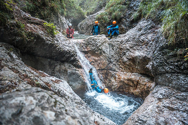 Geared up in neoprene suits and safety helmets, a Black male and his diverse friends embark on an outdoor canyoning adventure, sliding down rocky terrains.