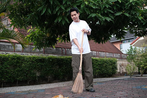 A man is sweeping the ground with a broom. The scene is outdoors and the man is wearing a white shirt