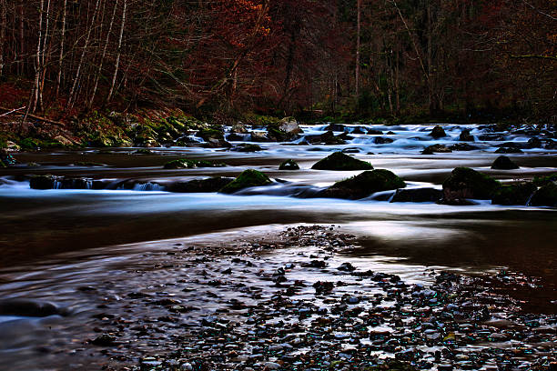 Close-up view of a rocky riverbed with large, moss-covered stones in the foreground. The silky, blurred water is the result of a long exposure, emphasizing the motion of the water and creating a moody, dramatic atmosphere.