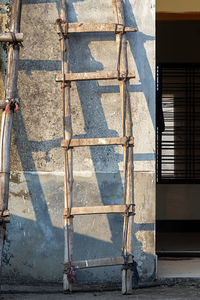 An old, worn handmade ladder crafted from rough-hewn bamboo and natural wood, leaning against the highly weathered, textured rooftop wall of a rustic building. Rural architecture and decay.