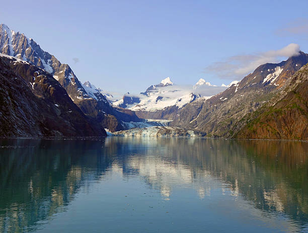 Towering snow-capped peaks rise above a crystal-clear fjord, their grandeur mirrored perfectly in the still waters below. At the heart of the scene, the brilliant blue ice of Johns Hopkins Glacier flows between rugged mountain walls, all beneath a radiant Alaskan sky. Captured in Glacier Bay National Park along Alaska’s Inside Passage, this breathtaking image showcases the untouched beauty and powerful serenity of one of the world’s most awe-inspiring landscapes. Ideal for lovers of wild places and dramatic natural scenes.
