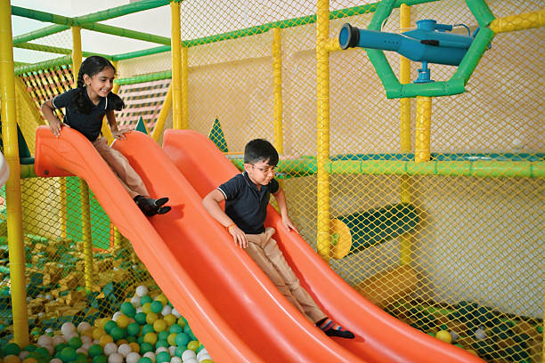 Primary school student glides down bright slide while friend waits below and classmates climb play structure behind. Childhood happiness and playful energy in a safe and engaging environment.