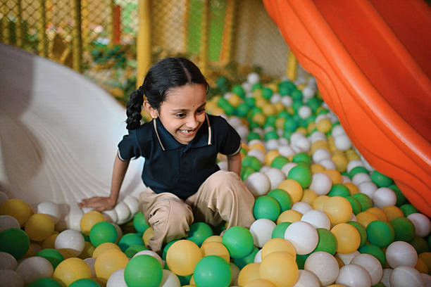 A cheerful primary school girl enjoys sliding into a vibrant indoor ball pit filled with yellow, green, and white plastic balls. Captured mid-laughter, she exudes joy and excitement as part of a fun playtime session. Carefree childhood moments, active play, and school-life balance. Early education, kids’ recreation, happiness, and playful learning environments.