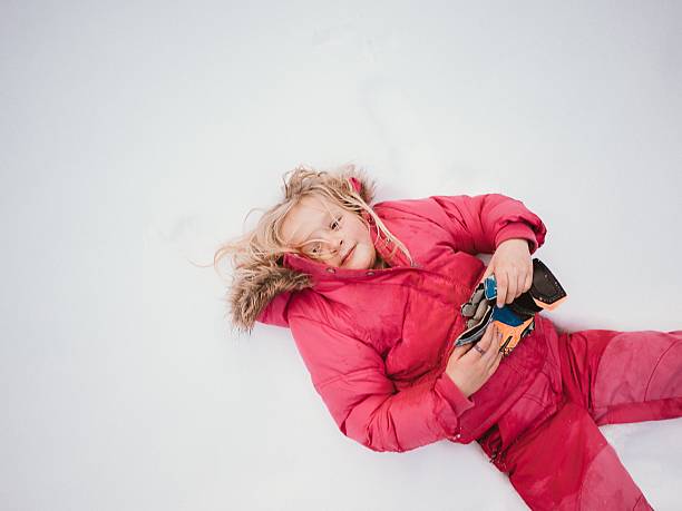 Child in pink winter attire lying on snow, holding ski gear. Relaxed expression reflects enjoyment of winter activities and play in a snowy outdoor setting