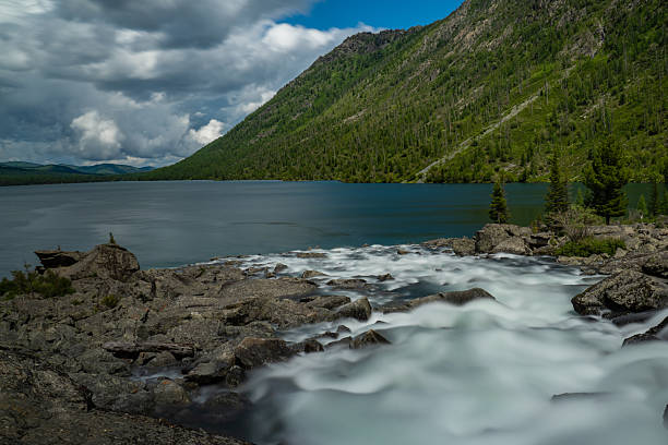 Scenic view of Lower Multinskoe Lake with deep blue clear water, river outflow with smooth long-exposure effect, rocky foreground, and forested green mountain slopes under dramatic clouds. Suitable for nature, travel, adventure, wilderness, and landscape themes, with ample copy space and untouched environment.