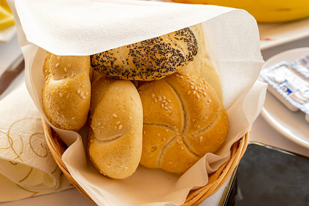 Assorted fresh round kaiser and sesame buns in a wicker bread basket with linen napkin on a restaurant breakfast table, closeup of golden crust bakery rolls for catering, brunch, lunch or dinner