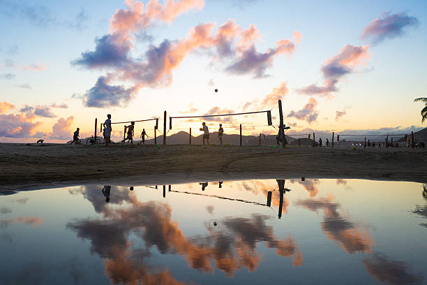Silhouettes playing beach volleyball at sunset in Santos, with colorful clouds reflected in a calm water puddle on the sand. Scenic and dynamic coastal scene.