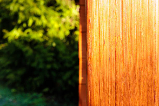 Horizontal photograph showing a warm wooden post on the right edge of the frame and softly blurred green foliage on the left. Strong sunlight reveals vertical grain lines and natural texture of the timber, while the out of focus garden creates a calm backdrop of rich color. The asymmetrical composition leaves generous copy space for text or graphics. This image is perfect for eco living concepts, outdoor lifestyle advertising, garden cabins, sustainable construction and real estate promotion.