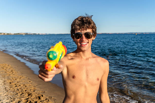 Young man smiling shirtless, aiming a water gun at camera while enjoying sunny beach fun by the ocean waves and sand