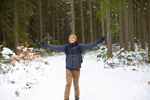 A happy man wearing a winter jacket, beanie and backpack walks along a snow-covered forest trail with his arms open wide. The coniferous forest and fresh snow create a joyful outdoor travel, freedom and winter adventure atmosphere