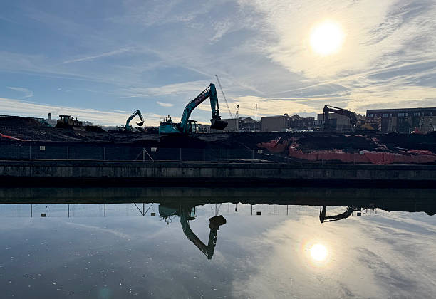 A construction site with excavators by the River Avon in the city of Bath, in Somerset UK. The calm water mirrors the machinery and the sky, creating a striking visual contrast between industrial activity and natural tranquility.