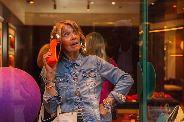 Mature woman holding an orange shoe to her ear while standing by a shop window