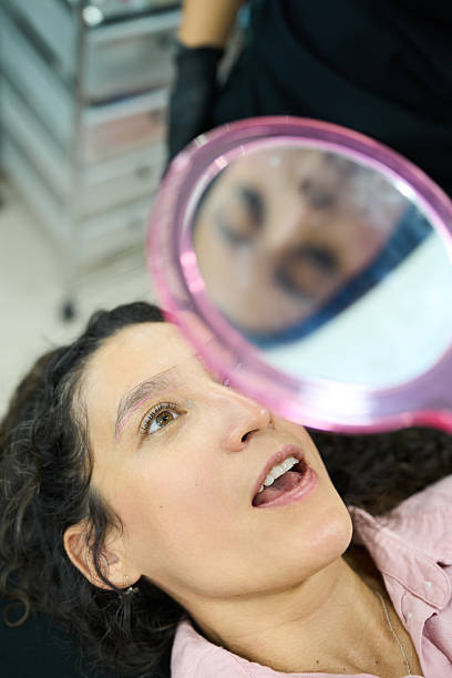 Young woman checking her newly tinted eyebrows in a small mirror at a salon, surprised and pleased with her fresh look and confidence