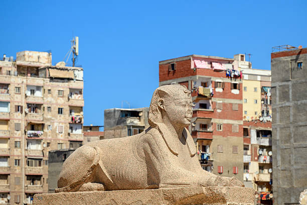 The ancient pink granite Sphinx located alongside Pompey's Pillar at the Serapeum Complex with the city skyline of Alexandria, Egypt in the distance.