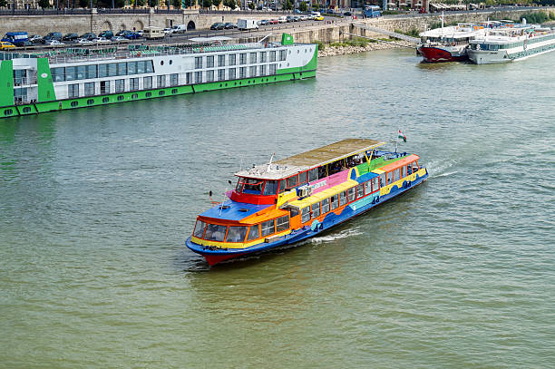 A vibrant, multi-colored passenger boat is cruising on the Danube River in Budapest, Hungary. The scene is an elevated view showing other docked river cruise ships and the Buda embankment and road traffic in the background on a sunny day.