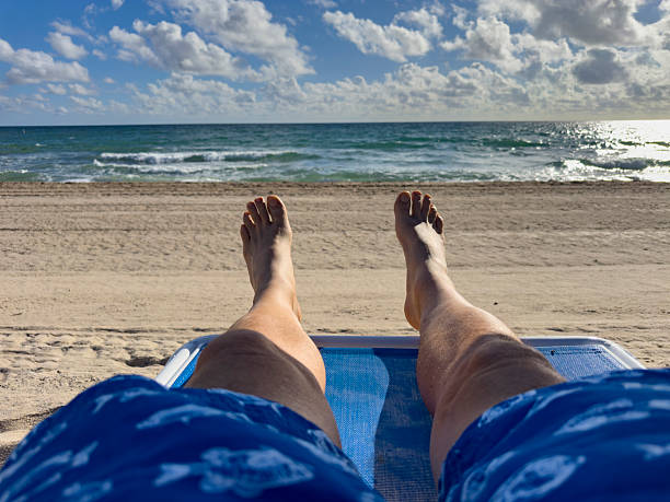 A first-person perspective of someone lounging on a beach chair, wearing blue swim shorts, and enjoying a sunny day at Lauderdale-by-the-Sea. The calm waves, soft sandy shore, and scattered clouds across the blue sky evoke a perfect moment of relaxation and coastal leisure.