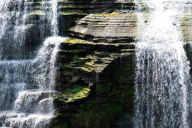 Water flows gracefully over rocky cliffs, creating several cascades in a peaceful natural landscape. Lush green plants thrive around the waterfall. Sunlight illuminates the scene, showcasing the water's movement and the texture of the stone.