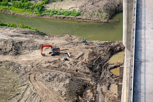 Heavy machinery is at work on the riverbank, clearing land near a dam. An excavator and other equipment are positioned among piles of dirt, while workers can be seen overseeing the operation.