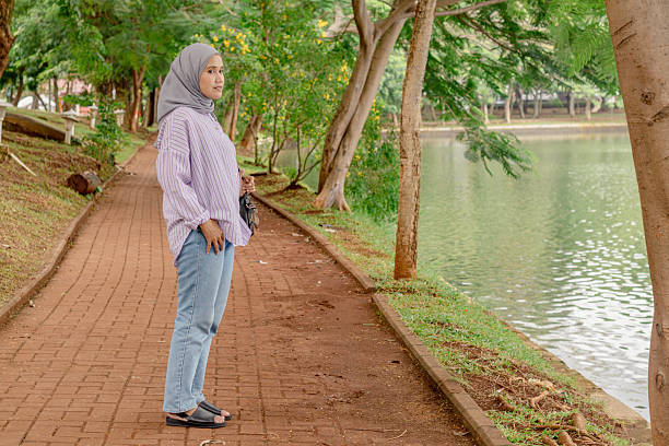 A young Asian Muslim woman standing on a walking path by a tree-lined lake