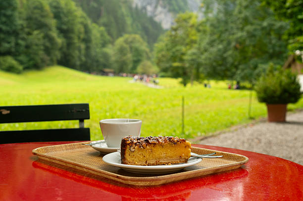 A delicious slice of nut and apple cake, possibly a Nusstorte, and a cup of coffee are placed on a red outdoor table. The background is a stunning, blurred view of the green Lauterbrunnen Valley.