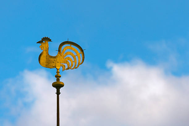 Close-up of golden weather vane atop a tiled roof, pointing against a clear, vibrant blue sky background.