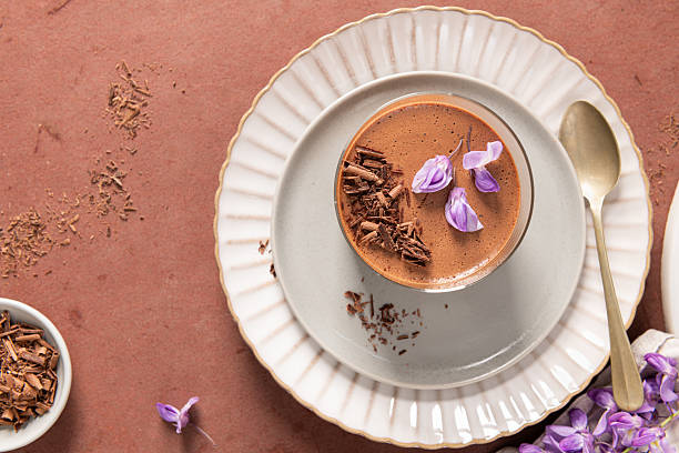 istock An overhead view features one creamy chocolate mousse in a glass, styled on plates with a spoon, a bowl of shavings, and purple flowers on a rustic surface. 2248672790