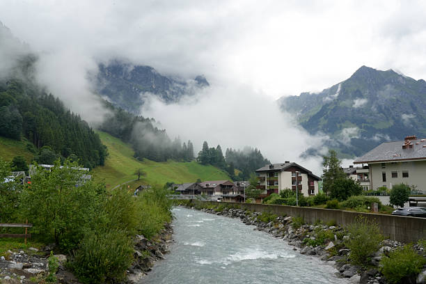 Mountain river flowing through Engelberg with alpine houses, green slopes and low clouds surrounding the steep mountain peaks. Photo taken on July 19, 2025.