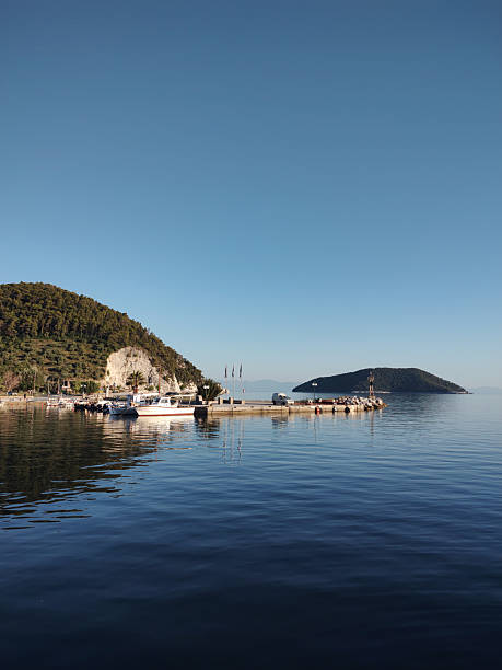 Scenic coastal view featuring a calm turquoise sea, pebble beach, anchored motorboats, and a water inflatable park in the distance. The coastline is lined with lush trees and hillside homes, all beneath a vibrant clear sky. Perfect for themes such as beach tourism, European summer destinations, Mediterranean coast, tranquil seascapes, vacation travel, nature backgrounds, and outdoor recreation.