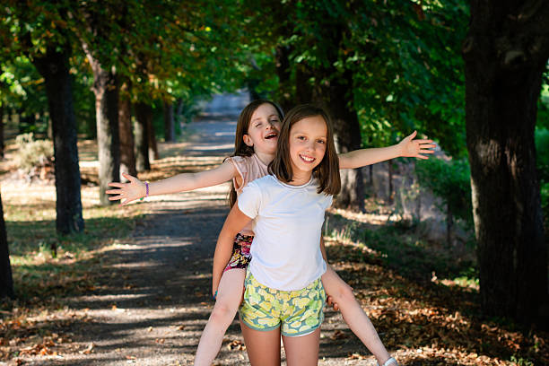 Two cheerful girls enjoying a piggyback ride on a tree-lined path, spreading arms like wings and laughing in a park