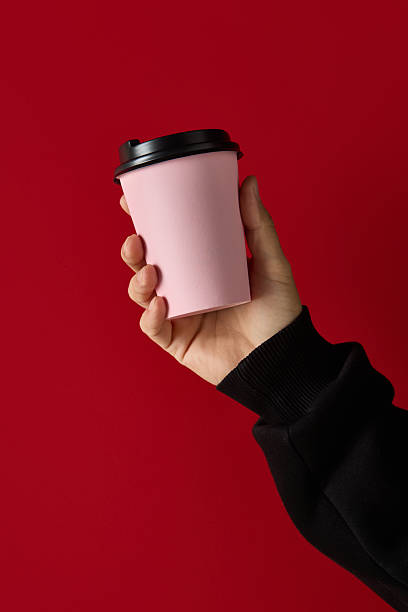 istock Hand holding pink takeaway coffee cup with black lid on red background for cafe packaging 2248679707