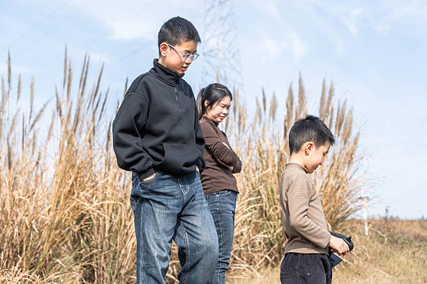 A family consisting of an adult female, a boy, and a young male are walking outdoors in a field with tall grass on a sunny day. They are dressed casually and seem to be enjoying a leisurely time together in nature