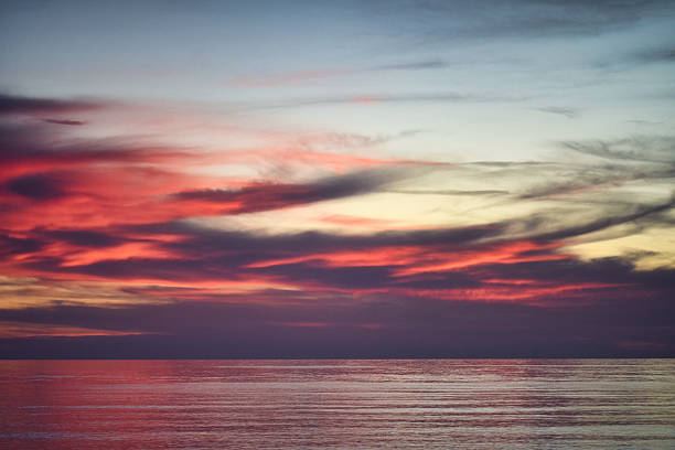 A view of vivid red, purple, and yellow sunset clouds above a calm sea. The layered cloud formations create strong color contrasts, with reflections visible on the water surface.