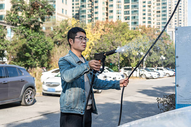 A man wearing a denim jacket is using a high-pressure water gun to clean something outdoors in a parking lot with several cars and buildings in the background