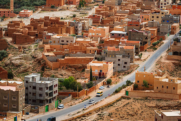 Aerial view of a Moroccan town with sun-baked clay houses, narrow streets, and a winding road. Warm earth tones, compact neighborhoods, and a traditional urban desert atmosphere.