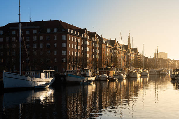 Warm evening light washes over the quiet canal in Copenhagen, illuminating the moored boats and historic brick buildings, creating a peaceful and picturesque waterfront scene.