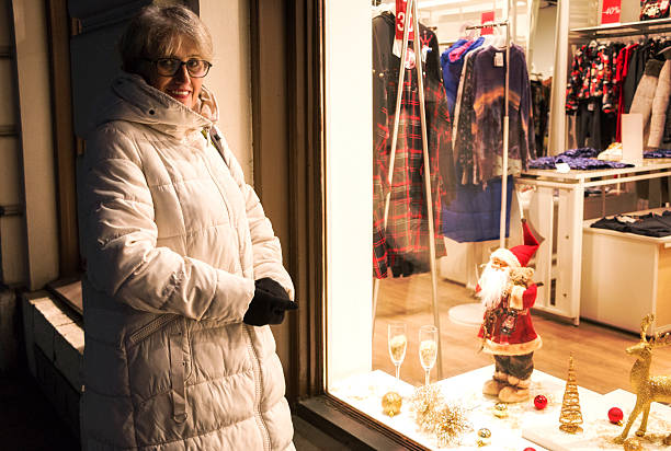 Senior  woman in a white coat looking at a shop window. She is looking for Christmas gift